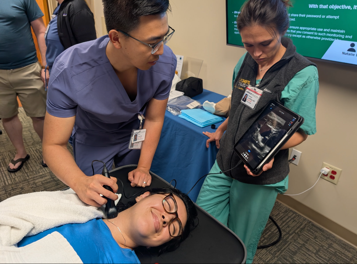 Three residents and medical students learning to use the butterfly ultrasound, as one student lays on a clinic bed.