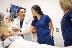 A clinical instructor in a white coat talks to two students in blue scrubs as they look at a simulation patient model in a hospital bed with a screen behind
