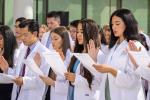 Students wearing white coats stand side-by-side with right palms up and left hands holding piece of paper