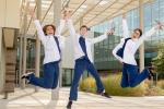 Three students wearing white coats and blue scrubs jump in the air with arms overhead