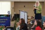 Woman stands on stage holding microphone looking down at those seated at table next to stage; monitor reads “Vision Statements 2023 Cohort”