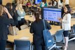 Woman stands at TV monitor showing scholarly project and talks to onlookers surrounding table