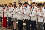 A group of students wearing white coats stand in a row, holding papers and raising their right hands as they take an oath during a ceremony.