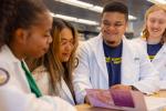 Four medical students in white lab coats that read “UC Davis School of Medicine” converse in front of a book at a library table.