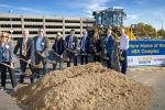 Pile of dirt with 9 individuals holding up shovels loaded with dirt, yellow front loader parked to the left