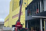 A red crane hoists a yellow wall pane with blue ribbon, watched by people in neon orange vests 