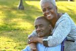 Black woman embraces husband from behind his shoulders while he sits in a wheelchair outdoors
