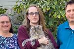 A family of three, mom, dad and teenage son, stand outside in a garden, while the teenage son holds a fluffy gray cat.