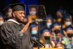 Graduate in cap and gown speaks at a podium, raising his hand beside a teleprompter, with masked graduates seated behind him.