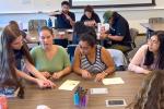 A student leader in blue shirt leans into a table to demonstrate beading techniques to four fellow students 