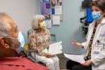 Geriatrician, right, wearing mask and white lab coat and holding a paper looks at male patient in exam room, while patient’s wife looks at doctor hold