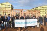 A group of about 40 people, most wearing coats and many wearing hats, stand together at a homebuilding site, behind a large white banner