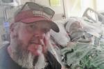 Bearded man with sunglasses and visor on top of his head sits next to hospital bed where he son lies in hospital gown and ventilator