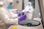 Person wearing blue mask and white PPE suit places vile in machine near another person in a blue mask and white PPE suit 