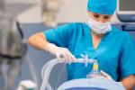 Nurse anesthetist in scrubs holding mask with tube over mouth of patient lying on the operating room table