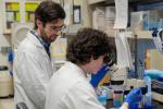 A person with dark curly hair seated at a lab bench looks into a microscope; another person with a beard and glasses wearing a white lab coat looks on
