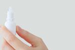 A hand holds a white eyedrop bottle against a light grey background. 