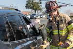 Firefighter in full gear stands outside a white truck holding fire boot while driver places dollar bill into boot   
