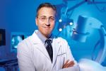 A middle-aged man in glasses and wearing a white physician coat stands with his arms crossed in front of some medical equipment. 