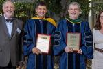 Three men and one woman stand in front of a shady tree; the two men in the middle are in academic robes and hold placards. 