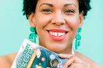 Woman with dark curly hair, green earrings and wearing a white dress holds a paperback book with the title, “Magnificent Makers.”