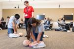 A woman in red polo shirt stands and gestures as two teenaged students, a boy and a girl, practice CPR on manikins on the floor 