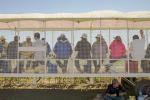 A row of farmworkers are shown sitting on an elevated platform covered with a cloth to provide shade. 
