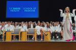 A medical student pumps her arms in celebration on stage at the white coat ceremony with fellow students seated in the background