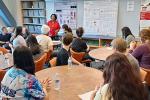 A woman in a red blouse stands at the front of a classroom before about 15 high school students seated around tables