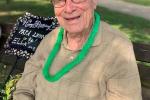 96-year-old man sits on a park bench, wearing a tan hat, glasses and a green lei around his neck. 