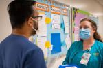 Two nurses wearing masks stand talking in front of bulletin board