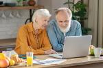Older woman sits next to older man both looking at computer screen in front of them on kitchen table