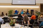 Three women sit on stage in front of room speaking into microphone while attendees sit at tables listening.