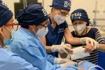 A woman wearing a blue surgical hat uses her hands to guide a woman putting a white drape on a manikin. 