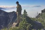 Firefighter stands on mountain range with smoke in the distance behind him