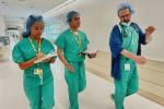 A teenage girl and a teenage boy, in green scrubs, take notes as they walk through a hospital hallway led by a doctor in scrubs