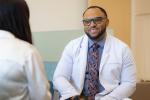 NP Chris Hall wears white coat, smiling and sitting across from patient whose back you see