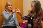 A woman with blond hair in a ponytail, wearing a blue long-sleeved top holds her right arm out and touches fingers with a gray-haired physician