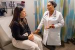 A doctor in a white coat, right, gestures with her left hand while speaking with a patient who is seated on an exam room chair