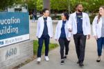Four students wearing white lab coats walking by blue sign that says Education Building