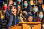 School of Medicine Student Speaker Rene Phan wearing graduation cap and gown takes selfie with her phone at the podium before her speech