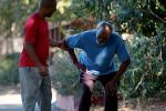 younger man in red shirt on left holds shoulder of older man in blue shirt who is bent over clutching thigh during walk outside