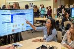 Student sitting at table points at research poster displayed on TV monitor