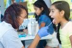 During a free clinic at the local park, a female clinician uses a tongue depressor to check preteen girl's throat. The clinician wears protective mask