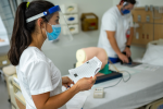 Nursing student wearing mask holds paperwork while other nursing student works on manikin in hospital bed in background