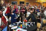 Docenas de hombres y mujeres parados en una sala de conferencia grande mueven sus brazos durante un ejercicio de grupo.