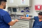 A man in blue polo shirt and baseball cap extends right arm on a counter so a student can take his blood pressure reading