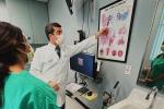 A male doctor in white coat speaks with a female teen in green scrubs in front of “Ear, Nose, Throat” poster