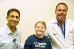A girl in a blue UC Davis Health Neurological Surgery shirt is flanked on the left and right by male doctors wearing badges.