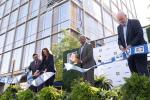 Four adults cut a ribbon on a stage in front of a large glass building. 